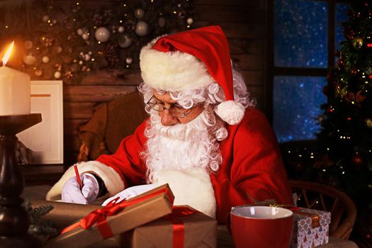 Portrait of happy Santa Claus sitting at his room at home near Christmas tree and answering Christmas letters