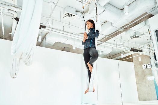 Ariel yoga. Young caucasian woman in sportswear practicing fly yoga in studio, standing on the one leg in white hammock and meditating, full length. Wellness and healthy lifestyle