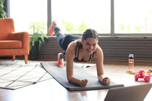 Beautiful fitness woman doing a plank exercise watching online tutorials on laptop, training in living room. Healthy lifestyle. Girl goes in for sports at home.