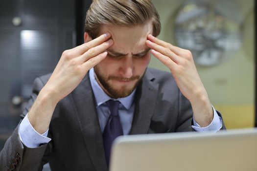 Image of tired young business man in suit keeping face in hands while sitting in the office