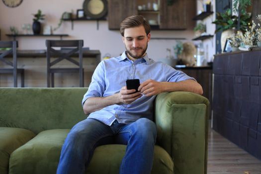 Attractive young man relaxing on a couch at home and using mobile phone for cheking social nets