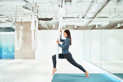 Antigravity. Young caucasian fitness woman in sportswear practicing fly yoga in beautiful bright studio. Sport, wellness and healthy lifestyle, aerial yoga concept