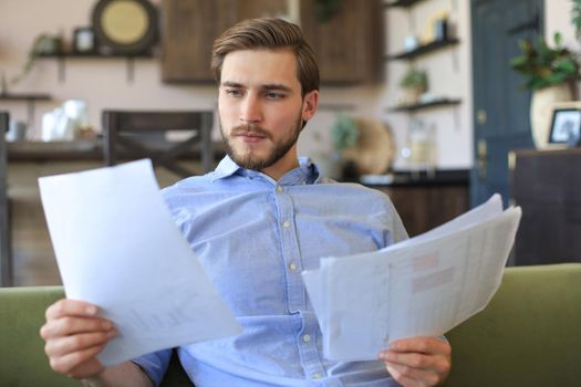 Concentrated young freelancer businessman sitting on sofa with laptop and examining documents