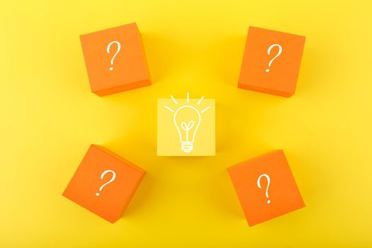 Concept of idea, creativity, start up or brainstorming. Flat lay with light bulb drawn on yellow cube and question signs on orange cubes against yellow background