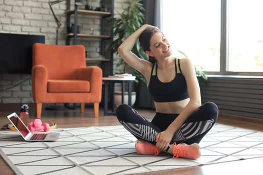 Fitness beautiful slim woman is sitting on the floor with dumbbells and bottle of water using laptop at home in the living room. Stay at home activities