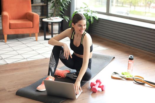 Beautiful slim sporty woman in sportswear is sitting on the floor with dumbbells and bottle of water and is using a laptop at home in the living room. Healthy lifestyle. Stay at home activities