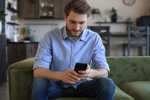 Attractive young man relaxing on a couch at home and using mobile phone for cheking social nets