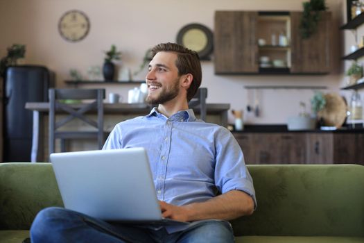 Happy young freelancer businessman sitting on sofa with laptop, working remotely online at home