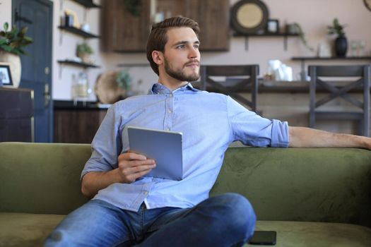 Smiling handsome man holding and using tablet with social networks at home, happy freelancer making online shopping, sitting on cozy sofa at living room