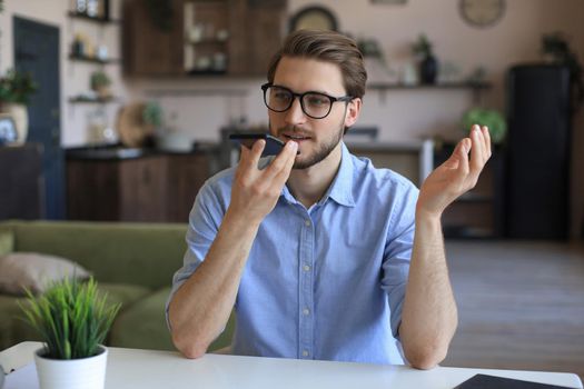 Confident business man in glasses holding smartphone near mouth for recording voice message or activating digital assistant