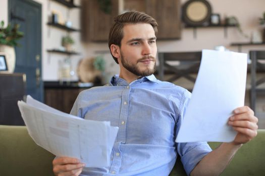 Concentrated young freelancer businessman sitting on sofa with laptop and examining documents