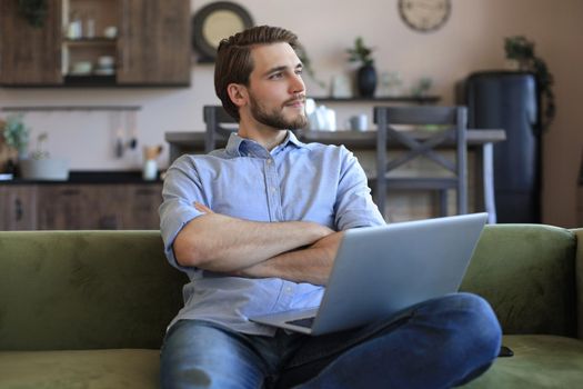 Concentrated young freelancer businessman sitting on sofa with laptop, working remotely online at home