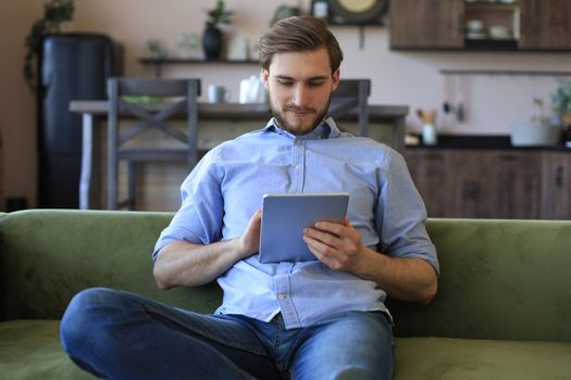 Smiling handsome man holding and using tablet with social networks at home, happy freelancer making online shopping, sitting on cozy sofa at living room