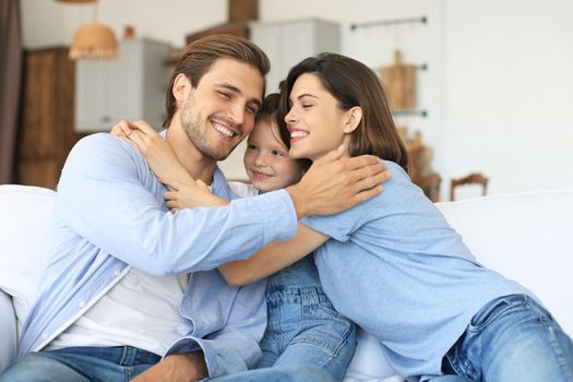 Positive friendly young parents with smiling little daughter sitting on sofa together while relaxing at home on weekend