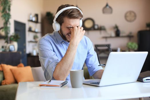 Unhappy frustrated young male holding head by hands sitting with laptop behind desk at home