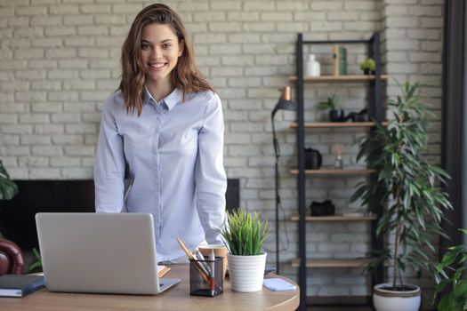 Confident young businesswoman with a friendly smile standing behind her desk in a home office looking at the camera