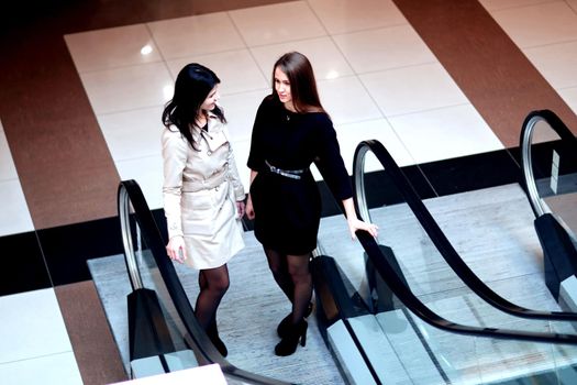 two business women talking standing on an escalator in a business center