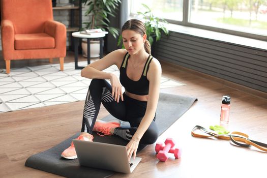 Beautiful slim sporty woman in sportswear is sitting on the floor with dumbbells and bottle of water and is using a laptop at home in the living room. Healthy lifestyle. Stay at home activities