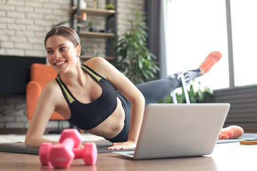 Fitness beautiful slim woman doing side plank with resistance band and watching online tutorials on laptop, training in living room. Stay at home activities.