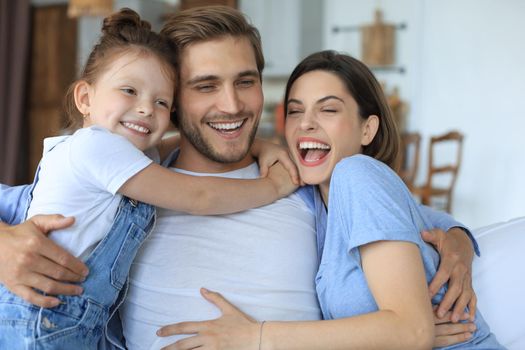 Positive friendly young parents with smiling little daughter sitting on sofa together while relaxing at home on weekend