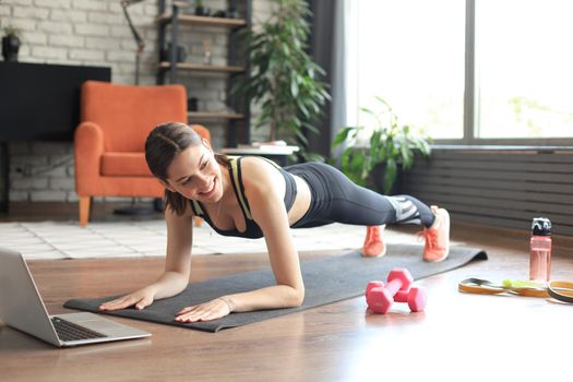 Fitness woman doing plank and watching online tutorials on laptop, training in living room. Stay at home activities. Top view