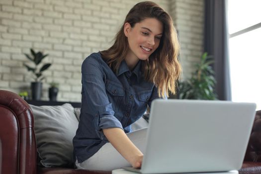 Smiling young woman sitting on sofa with laptop computer and chating with friends