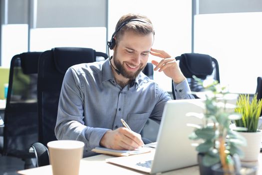 Smiling male call-center operator with headphones sitting at modern office, consulting online information in a laptop, looking up information in a file in order to be of assistance to the client