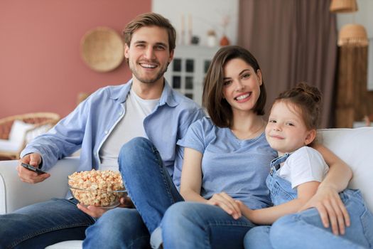 Happy family with child sitting on sofa watching tv and eating popcorn, young parents embracing daughter relaxing on couch together