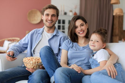 Happy family with child sitting on sofa watching tv and eating popcorn, young parents embracing daughter relaxing on couch together