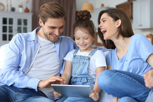 Positive friendly young parents with smiling little daughter sitting on sofa together answering video call on digital tablet while relaxing at home on weekend