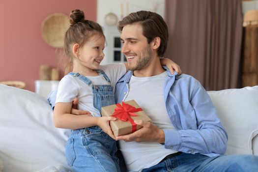 Cute kid daughter make surprise to daddy, little girl presents gift box to father sit on sofa. Father's day