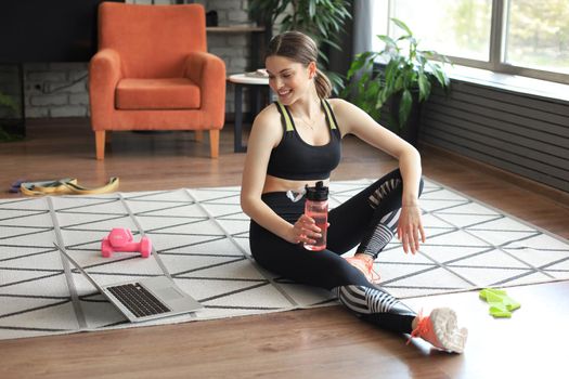 Fitness beautiful slim woman is sitting on the floor with dumbbells and bottle of water using laptop at home in the living room. Stay at home activities