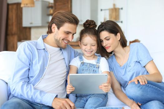 Positive friendly young parents with smiling little daughter sitting on sofa together answering video call on digital tablet while relaxing at home on weekend