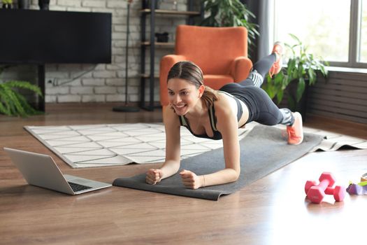 Beautiful fitness woman doing a plank exercise watching online tutorials on laptop, training in living room. Healthy lifestyle. Girl goes in for sports at home.