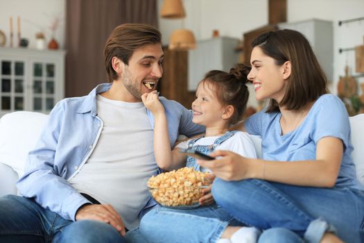 Happy family with child sitting on sofa watching tv and eating popcorn, young parents embracing daughter relaxing on couch together