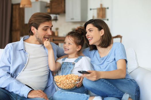 Happy family with child sitting on sofa watching tv and eating popcorn, young parents embracing daughter relaxing on couch together