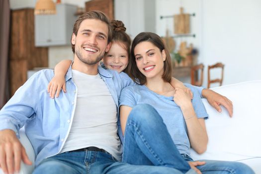 Positive friendly young parents with smiling little daughter sitting on sofa together while relaxing at home on weekend