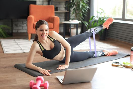 Fitness beautiful slim woman doing side plank with resistance band and watching online tutorials on laptop, training in living room. Stay at home activities.