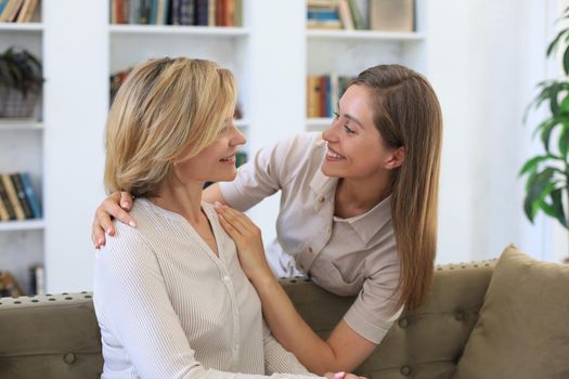 Beautiful middle aged mom and her adult daughter are hugging and smiling while sitting on couch at home