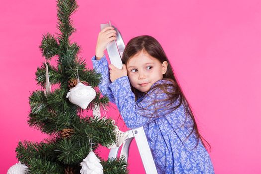 Children, holidays and christmas concept - little girl decorating christmas tree on pink background.