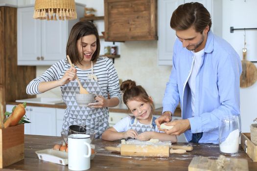 Cute little girl and her parents are having fun while cooking in kitchen at home together