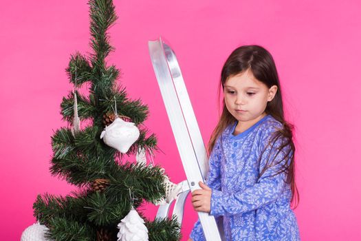 Children, holidays and christmas concept - little girl decorating christmas tree on pink background.