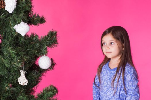 Christmas, childhood and people concept - little girl look at the christmas tree over pink background.