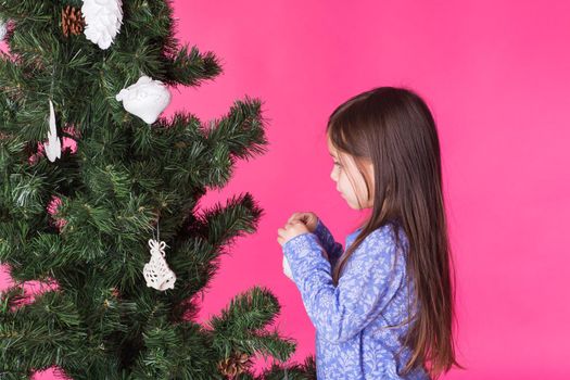 Little girl decorating christmas tree on pink background.
