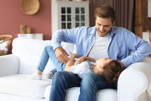 Smiling young father lying on couch at living room and play with happy little daughter