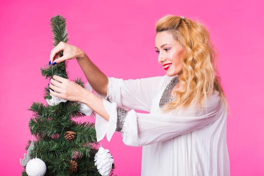 Christmas, holidays and people concept - young happy blonde woman decorating christmas tree on pink background.