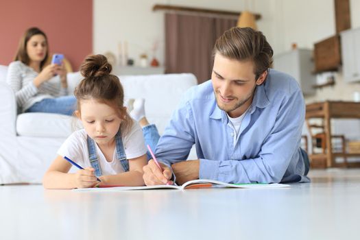Happy father smilling daughter lying on warm floor enjoying creative activity, drawing pencils coloring pictures in albums, mother resting on couch, family spend free time together