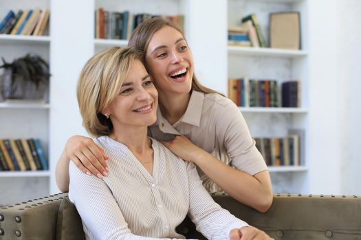 Beautiful middle aged mom and her adult daughter are hugging and smiling while sitting on couch at home