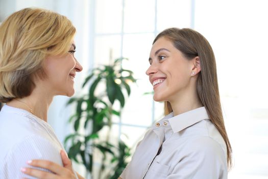 Cheerful young woman is embracing her middle aged mother in living room