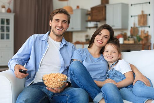 Happy family with child sitting on sofa watching tv and eating popcorn, young parents embracing daughter relaxing on couch together